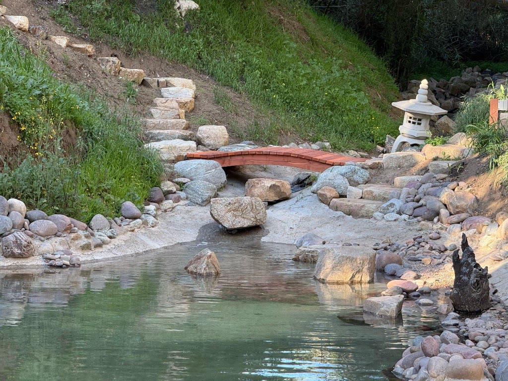 Cedar arched footbridge over a stone-lined pond channel with stone steps and a lantern